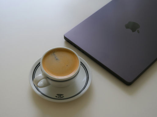 A white cup of espresso on a saucer beside a closed laptop on a light surface. The scene is simple and minimalist.