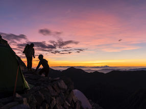 Group backpacking trip at sunrise on a mountain slope silhouette against a vibrant pink and orange sky, with tents nearby and distant peaks visible.