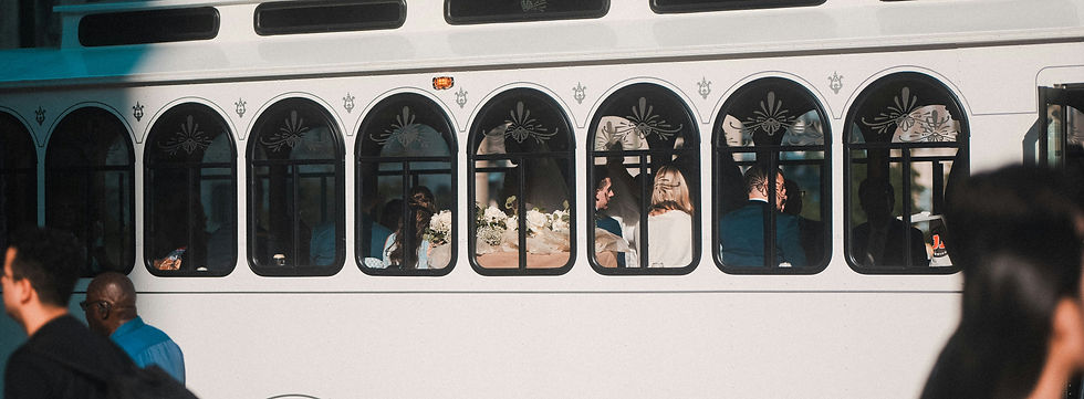 Passengers sit inside a white arched-window trolley decorated with flowers, while people walk past outside in sunlight.