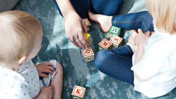 Children sitting on a blue rug playing with colorful alphabet blocks during a pediatric speech therapy session, while an adult hand arranges letters to support learning.