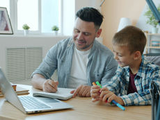 A man and boy smile while working on a notebook at a desk. A laptop, plants, and books are nearby. Bright, cozy room with large windows.