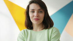 Woman with short brown hair in a light green sweater smiles slightly against a colorful geometric background in yellow, blue, and orange.