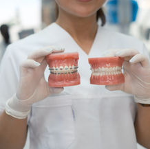 A dental professional holds two dental molds, one with braces, in a clinic. They wear white gloves and a uniform, with a blurred background.
