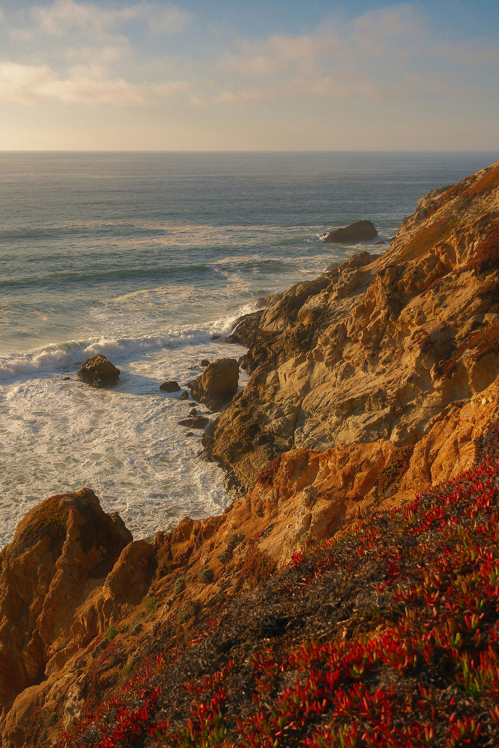 Sunlit rocky coastline with waves crashing against the cliffs. Red vegetation covers the foreground, under a partly cloudy blue sky.