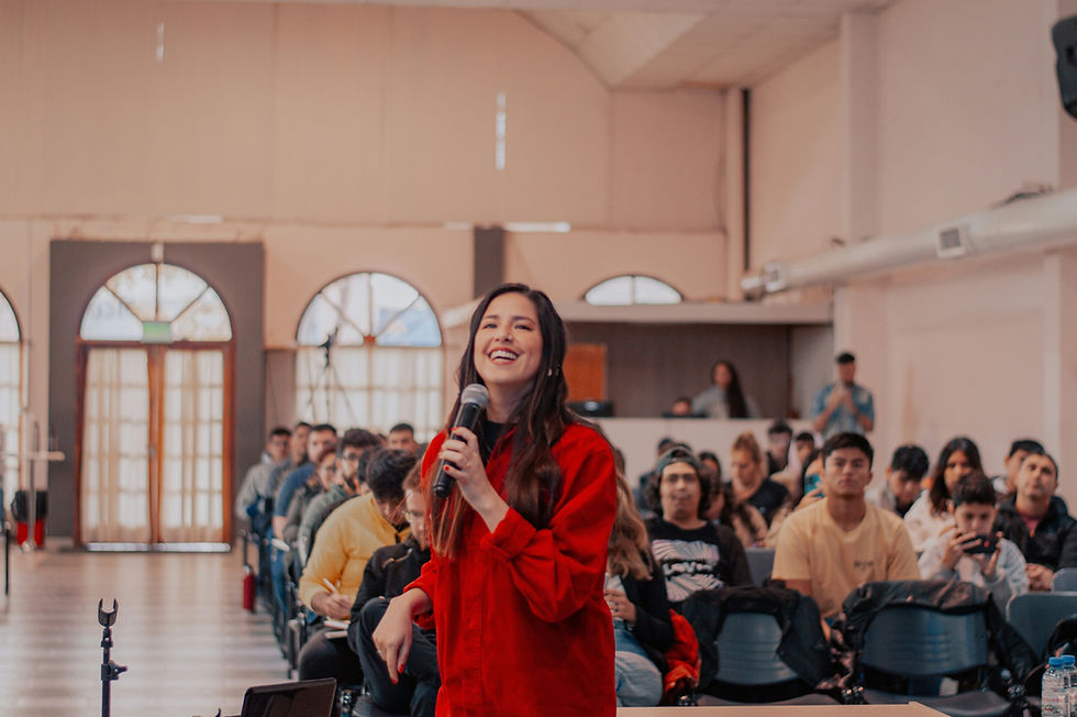 Woman in red shirt speaking to a seated audience in a hall, smiling, holding a microphone. Bright atmosphere, arched windows in background.