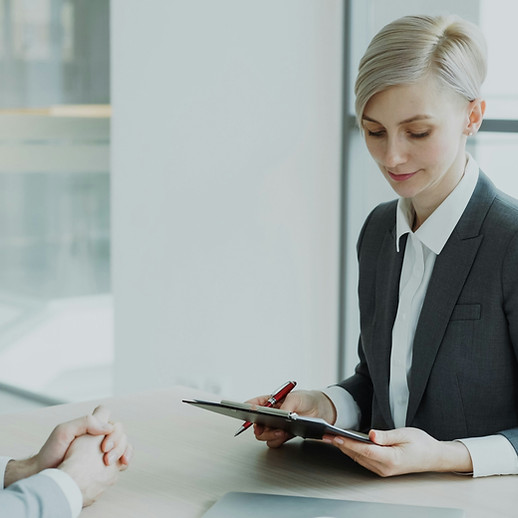 Two professionals in suits meeting at a desk, woman taking notes on clipboard