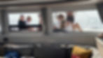 People relax on a boat deck viewed through a window. Inside, a sofa with colorful pillows is visible. Sunny and serene atmosphere.