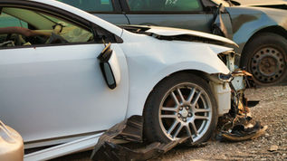 Voiture blanche accidentée, pare-chocs arraché et capot ouvert, près d’une autre voiture cabossée; graviers, notes sur la vitre.
