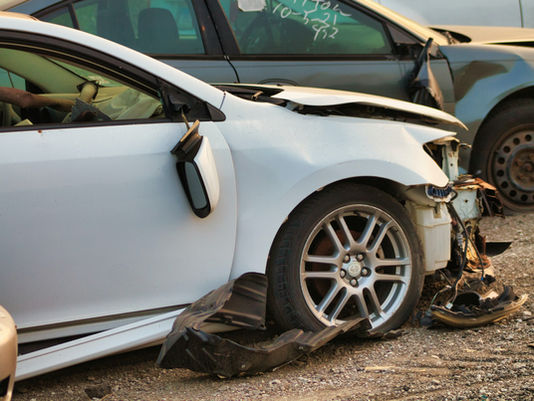 Voiture blanche accidentée, pare-chocs arraché et capot ouvert, près d’une autre voiture cabossée; graviers, notes sur la vitre.