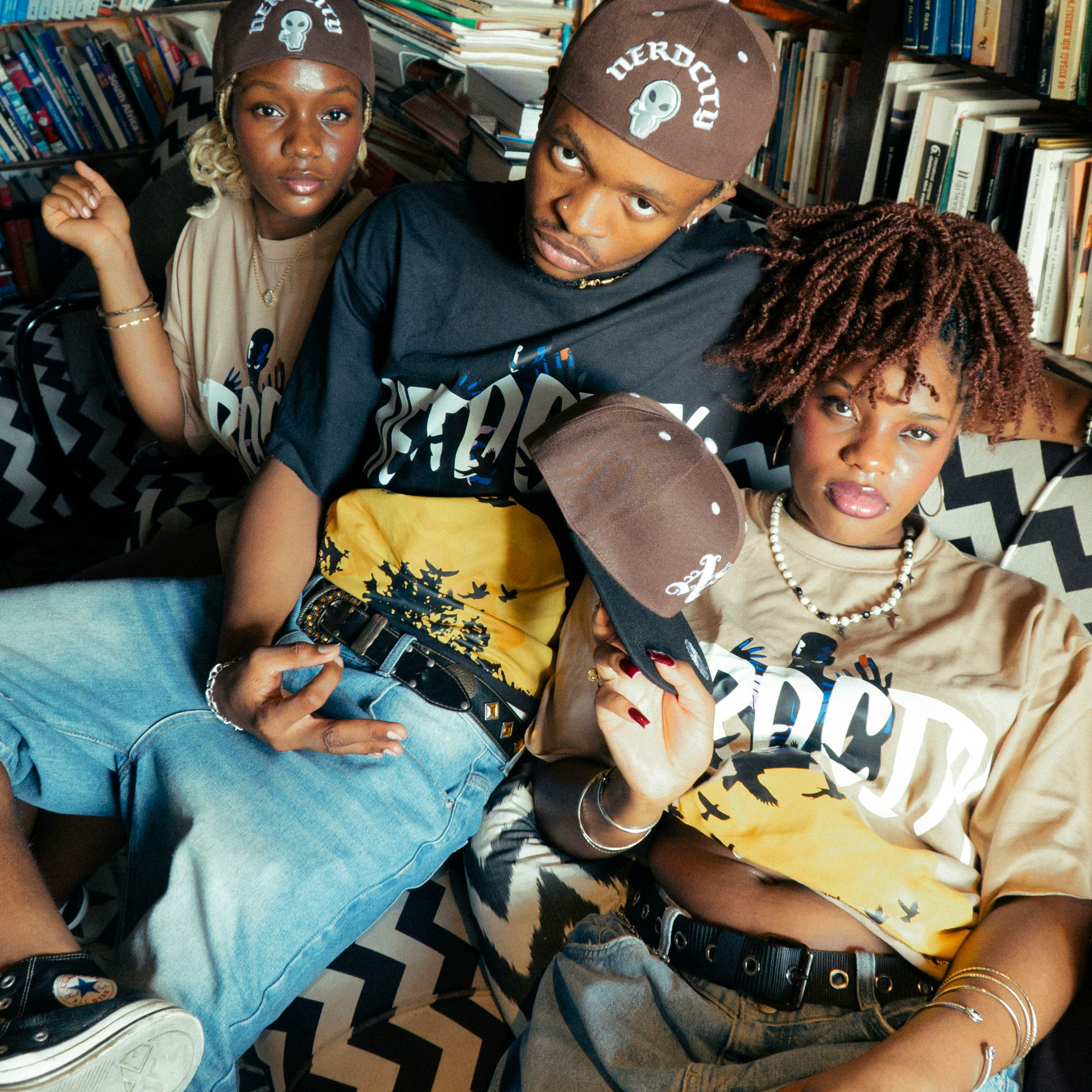Three people in casual wear sit on a patterned couch in a library, wearing matching caps. They convey a relaxed, stylish vibe.