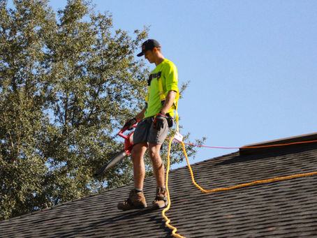 Worker in a neon shirt, using a leaf blower, stands on a roof with safety harness and ropes. Tree and blue sky in the background.