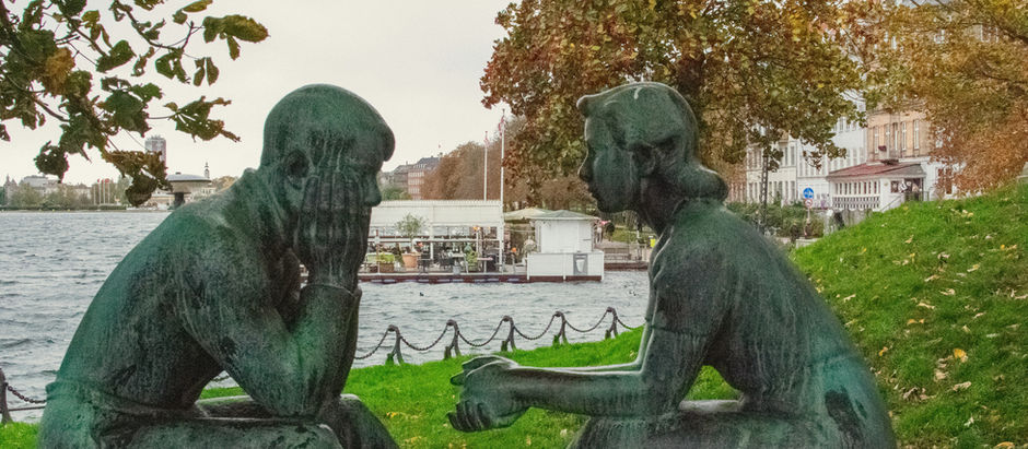 Two bronze statues seated by a waterfront, facing each other in conversation, symbolising intimacy, vulnerability, and emotional reflection in relationships.