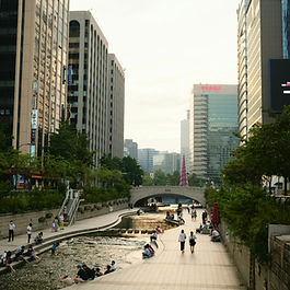 Transforming a floodwater system: how Seoul turned Cheonggyecheon Stream into an urban renewal success story Seoul, South Korea’s bustling capital, is renowned for its rapid development and modern skyline. However, beneath its high-rise buildings lies a story of urban transformation that has reshaped the city’s landscape. Cheonggyecheon Stream, once buried beneath an elevated highway, has been revitalised into a vibrant public space that enhances both the city’s aesthetics and liveability. The project has served as a model for cities worldwide, demonstrating the potential for urban waterways to support environmental sustainability, economic growth, and social well-being. This article explores how the restoration of Cheonggyecheon Stream revitalised Seoul and what lessons other global cities can draw from this ambitious initiative. The evolution of Cheonggyecheon Stream Cheonggyecheon Stream has played a significant role in Seoul’s history for over 600 years. Initially a natural waterway, it supported daily life and acted as a crucial flood management system. However, as the city expanded, the stream deteriorated into an open sewer, becoming a public health concern. During the 1960s and 1970s, as part of South Korea’s push for rapid urbanisation and modernisation, the stream was covered by concrete, and an elevated highway was constructed above it. This infrastructure was hailed at the time as a sign of progress, but by the late 20th century, it had become clear that prioritising road networks over public spaces came at a cost. The case for restoration By the late 1990s, public attitudes towards urban development in Seoul were shifting. Several infrastructure failures, including the tragic collapses of the Seongsu Bridge in 1994 and the Sampoong Department Store in 1995, raised concerns about the city’s ageing structures. Moreover, Seoul’s rapid urbanisation had led to increased congestion, pollution, and reduced green spaces. The government faced a choice—continue maintaining the deteriorating elevated highway or reimagine the space for a more sustainable future. In 2002, the newly elected mayor of Seoul, Lee Myung-bak, championed the restoration of Cheonggyecheon Stream as a key urban renewal project. The initiative aimed to improve air quality, reduce traffic congestion, create recreational spaces, and establish a more environmentally friendly cityscape. Despite concerns about potential traffic gridlock, the project moved forward with strong political backing and public support. The transformation process The restoration project, completed in just 27 months at a cost of 386 billion won (approximately £220 million), involved the removal of the elevated highway and the reconstruction of the stream. The revitalised Cheonggyecheon Stream now spans nearly 6 km through Seoul’s central districts, featuring pedestrian-friendly pathways, stepping stones, green spaces, and cultural installations. The project also incorporated sophisticated flood management infrastructure to handle extreme rainfall events. Since its reopening in 2005, Cheonggyecheon Stream has become a focal point for residents and visitors alike. The area is now 3.6°C cooler than surrounding streets, reducing the urban heat island effect. Improved air circulation has led to a noticeable decline in pollution, and the return of wildlife has restored biodiversity. Furthermore, the project has reinvigorated the local economy, with businesses and tourism flourishing around the revitalised space. Economic and social impact The restoration of Cheonggyecheon Stream has had a profound economic impact on Seoul. Property values in the area surged, with office rents increasing by 13% and land prices rising between 35% and 80% in redevelopment districts. The transformation of the stream also led to a shift in land use from industrial zones to commercial hubs, hotels, and cultural venues. Socially, the stream has provided much-needed public space in one of the world’s most densely populated cities. It serves as a cultural corridor, hosting year-round festivals, exhibitions, and performances. The Seoul Lantern Festival, an annual event featuring intricate paper lantern displays, has become a significant attraction, drawing millions of visitors. Additionally, the stream’s green spaces offer residents a tranquil retreat from the fast-paced urban environment, promoting well-being and outdoor activities. Challenges and criticisms Despite its success, the Cheonggyecheon restoration has not been without challenges. Unlike its historical counterpart, which flowed naturally, the modern stream relies on artificial water supply. Approximately 48,000 tonnes of water are pumped daily from the Han River and groundwater sources to maintain its flow, leading to concerns about sustainability and maintenance costs. The city spends 2.9 billion won annually on upkeep, which has raised debates about whether the project is truly an environmental success or simply an aesthetic improvement. Nevertheless, the overall benefits of the project—ranging from improved urban resilience to increased economic opportunities—have outweighed these concerns. The success of Cheonggyecheon has encouraged further urban renewal initiatives in Seoul, including the removal of 16 other elevated highways and the creation of more pedestrian-friendly spaces. Lessons for global cities The transformation of Cheonggyecheon Stream serves as an inspiring example for cities worldwide looking to revitalise their urban environments. Many global cities face similar challenges of ageing infrastructure, environmental degradation, and limited public spaces. The Seoul model demonstrates that reclaiming waterways and integrating them into urban planning can offer significant ecological, economic, and social benefits. From New York’s High Line, a repurposed railway turned into a linear park, to Utrecht’s conversion of a motorway back into a canal, cities are increasingly recognising the potential of transforming outdated infrastructure into valuable public spaces. By prioritising human-centred urban design, cities can enhance liveability, reduce pollution, and foster economic growth. Conclusion Seoul’s decision to restore Cheonggyecheon Stream has profoundly reshaped the city’s landscape, improving environmental quality, boosting the local economy, and providing a vibrant public space for residents and visitors. While challenges such as water management and maintenance costs remain, the project has demonstrated the immense value of integrating nature into urban spaces. As cities worldwide seek innovative ways to enhance sustainability and liveability, the success of Cheonggyecheon offers a compelling blueprint for the future. By reclaiming and revitalising waterways, urban centres can create healthier, more dynamic, and more attractive environments for generations to come. See https://www.georgejamesconsulting.com/ for more.