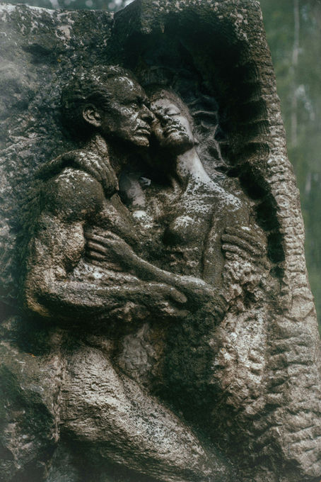 Stone sculpture of two intertwined figures embracing, with textured, weathered surfaces, set against a forested background. Their expressions suggest deep emotion.