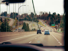 Cars drive on a multi-lane bridge road, surrounded by evergreen trees. Overcast sky with a green sign for Jackson Ave, 6th Ave, East, and Downtown.