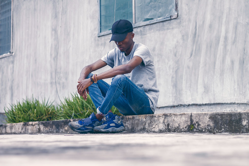 Man in cap and jeans sitting on concrete ledge, looking down thoughtfully. Gray wall and greenery in background, muted tones.