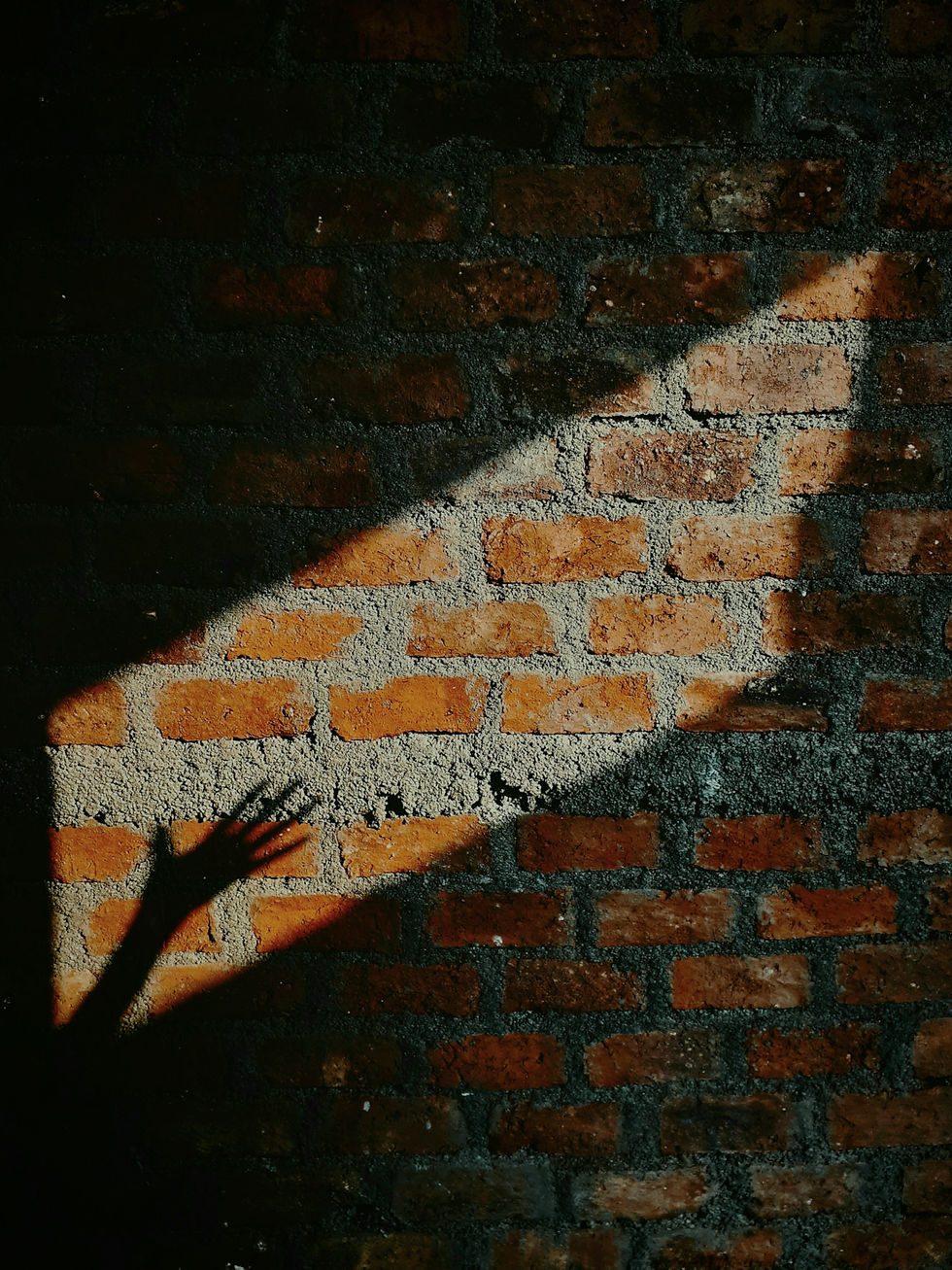 Shadow of a hand reaches across a sunlit red brick wall, casting a dramatic silhouette. The scene evokes a sense of mystery and intrigue.