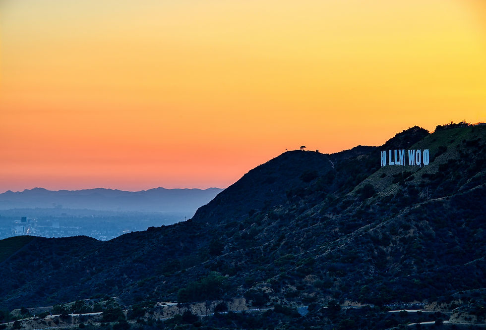 The sun sets over hills with the iconic "HOLLYWOOD" sign. The sky is a gradient of orange to yellow, casting a serene mood.