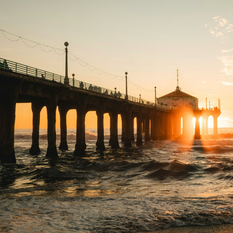 Sunset view of a pier over the ocean, with waves crashing. Warm orange and yellow hues illuminate the scene, creating a serene atmosphere.