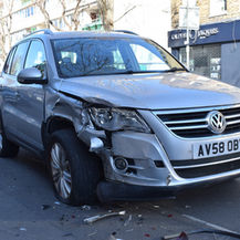 Damaged silver Volkswagen SUV with crushed front-left fender on a city street, license plate AV58 OBY, outside shops and parked cars