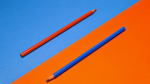 Two colored pencils, one orange and one blue, lie diagonally on a blue-and-orange split background.