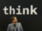 Man in glasses and blue blazer looks upward against a black background with large white text reading "think."