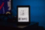E-reader displaying book list in a dimly lit room with a blue background. Nearby, a monitor shows abstract patterns. Mood is calm and tech-focused.